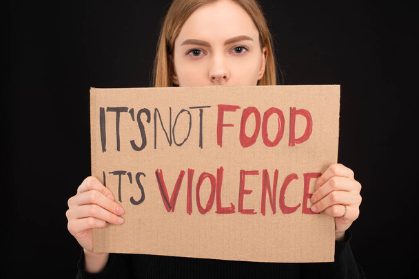 woman with obscure face holding cardboard sign with its not food its violence inscription isolated on black - 写真・画像