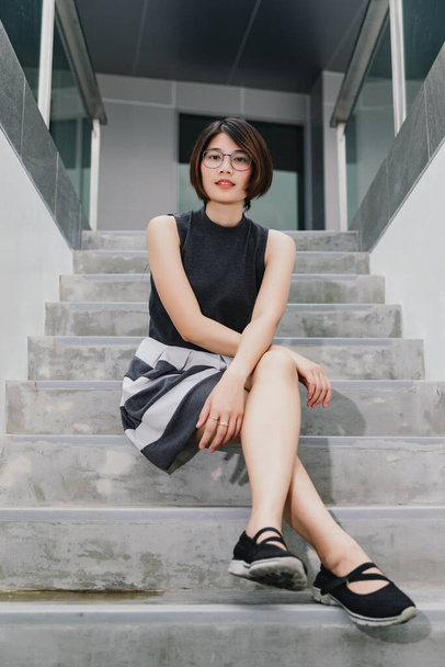 Retrato de asiático jovem cabelo curto bela mulher vestindo óculos e vestido cinza posando sentar pernas cruzadas nas escadas
 - Foto, Imagem