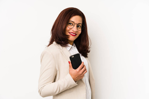 Středního věku latinská žena drží mobilní telefon izolované pohledy stranou s úsměvem, veselý a příjemný. - Fotografie, Obrázek