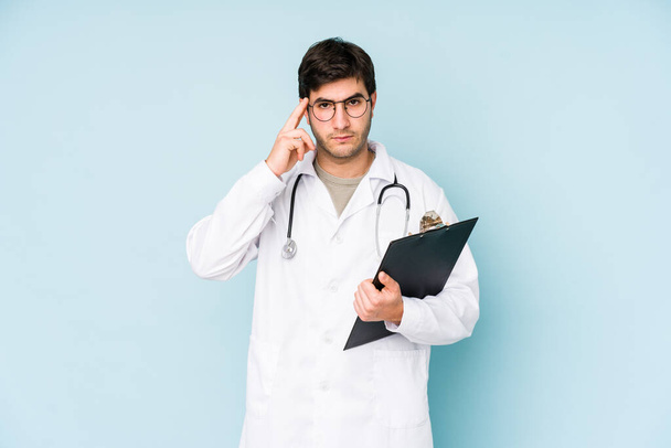 Молодой доктор, изолированный на синем фоне, указывающий пальцем на висок, думающий, сосредоточенный на задаче
. - Фото, изображение