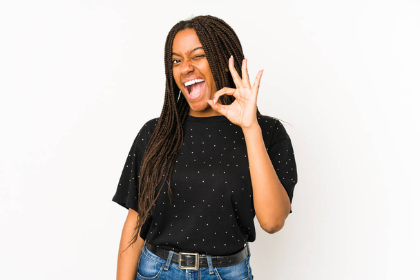 Молодая африканская американка, изолированная на белом фоне, подмигивает глазом и держит нормальный жест рукой.. - Фото, изображение