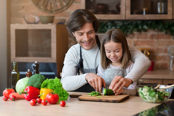 Padre premuroso che insegna alla sua figlioletta a cucinare, tagliando insieme il cetriolo
 - Foto, immagini