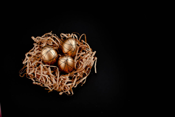Ostergold und weiß verzierte Eier im Nest auf schwarzem Hintergrund. Minimales Osterkonzept kopiert Platz für Text. Horizontale Ansicht von oben - Foto, Bild
