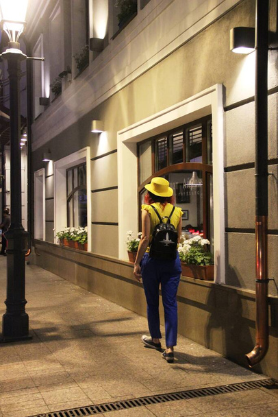 Eine Frau mit gelbem Hut geht weg, in der Ferne auf der nächtlichen Straße der Altstadt, schwarz-weißer Rucksack. Rückansicht einer Frau, die nachts auf der Straße in der Altstadt von Moskau spaziert - Foto, Bild