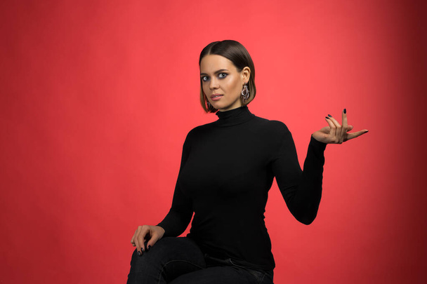 Beautiful emotional happy model with smokey makeup looking at camera and smiling, lady wearing black polo neck, presenting something by the finger, red background, concept of emotions with copy space - Photo, image