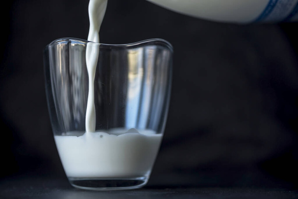Milch aus der Milchflasche in ein Glas auf schwarzem Hintergrund gießen, isoliert, Nahaufnahme - Foto, Bild