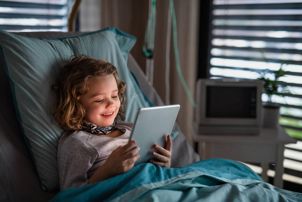 Набридла маленька дівчинка в ліжку в лікарні, використовуючи планшет, щоб пройти час
. - Фото, зображення