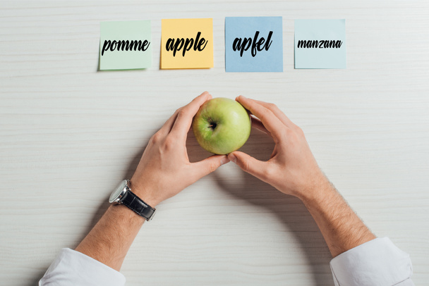 cropped view of businessman holding apple on table with sticky notes with apple translation - Photo, Image
