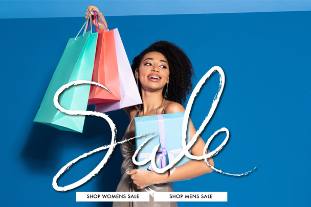 Gümüş elbiseli, hediye kutusu ve alışveriş çantaları taşıyan güzel Afro-Amerikan kadını mavi arka plana bakarken, satış illüstrasyonları - Fotoğraf, Görsel
