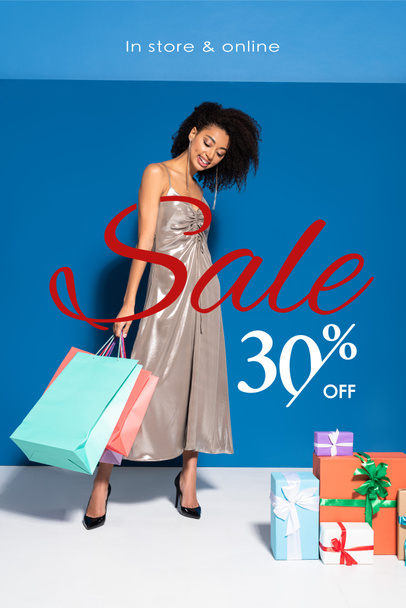 happy beautiful african american woman in silver dress holding shopping bags near gifts on blue background, 30 percent off sale illustration - Photo, Image