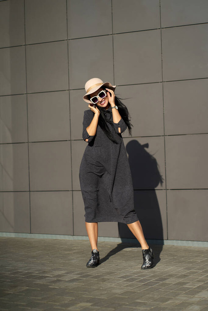 Naprosto. Šťastná roztomilá žena nosí mladé šedé šaty ve stylovém klobouku a módní sluneční brýle. Model v módní oblečení pózuje na městském šedém pozadí zdi a odvrací pohled.                              - Fotografie, Obrázek