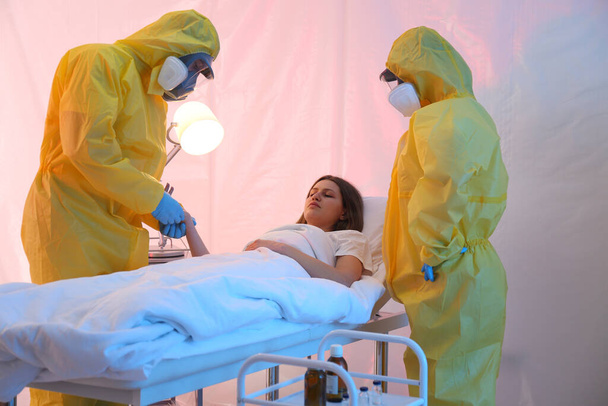 Paramedics wearing protective suits examining patient with virus in quarantine ward - 写真・画像