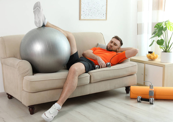 Lazy young man with sport equipment on sofa at home - Valokuva, kuva
