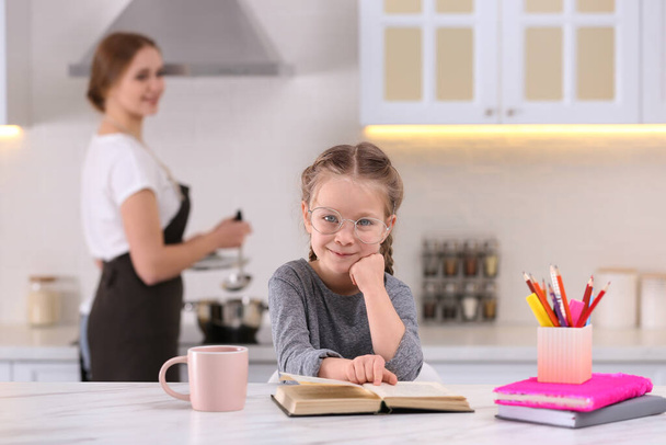 Little girl doing homework while mother cooking in kitchen - Zdjęcie, obraz