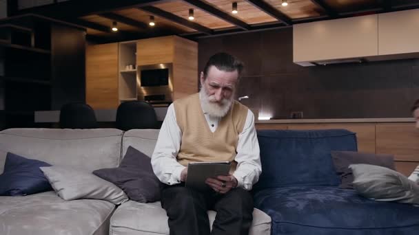 Family concept where serious bearded 80-aged man sitting on sofa working on tablet pc and his three cheerful teenage grandchildren running to him with huggs - Video