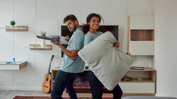 Having fun. Two young professional cleaners having fun while working together in the living room. Young caucasian man playing with handheld vacuum, afro american woman holding sofa pillow and smiling - Fotó, kép