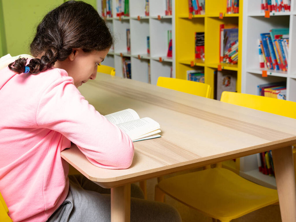 Genç kız öğrenci bir kitap okurken ya da okula ve eğitim konseptine dönerken bir masada ya da kütüphanede oturdu. - Fotoğraf, Görsel