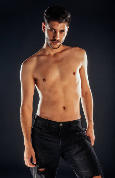 Modeporträt eines Oben-ohne-Mannes in zerrissenen Jeans vor schwarzem Hintergrund - Foto, Bild