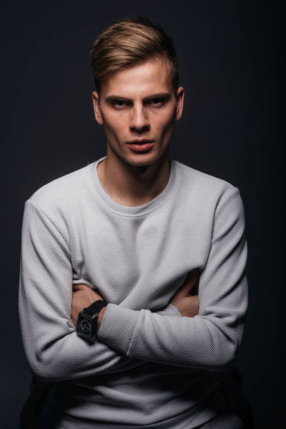 Informally ( casual ) dressed blonde young man with sharp jawline in his 20's posing in a studio in front of a black background while wearing a white sweater. - 写真・画像
