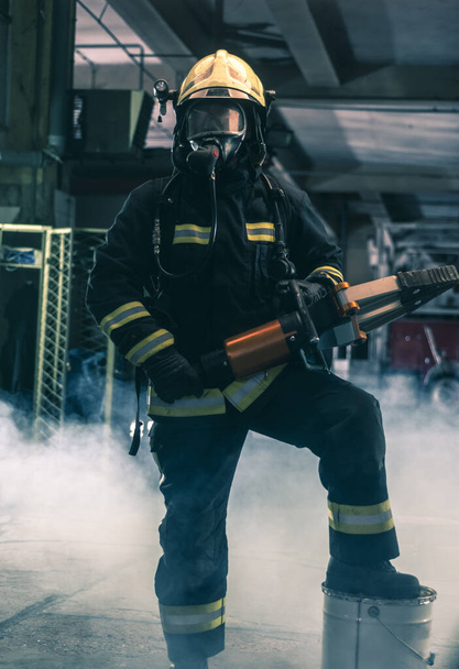 Портрет храброго пожарного, уверенно стоящего в полном защитном снаряжении, явки и шлеме. Темный фон с дымом и синим светом. - Фото, изображение