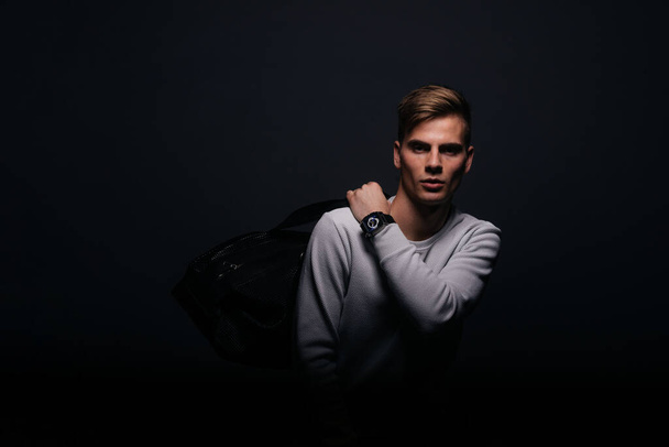 Nieformalnie (dorywczo) ubrany blondyn młody człowiek z ostrą szczęką w wieku 20 lat, pozujący w studio przed czarnym tłem, nosząc biały sweter i trzymając torbę czarnego mężczyzny. - Zdjęcie, obraz