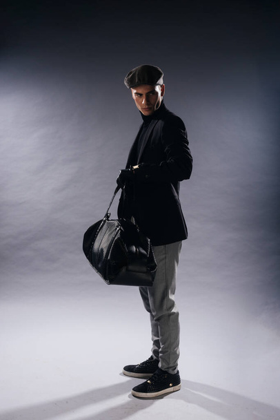 Muotokuva nuori mies terävä leuka yllään newsboy lippis, tilalla mies laukku poseeraa studiossa valkoisella pohjalla. - Valokuva, kuva