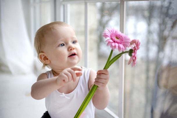 1 little blonde girl with beautiful blue eyes sitting by the window with pink flowers, portrait of a cute baby - 写真・画像