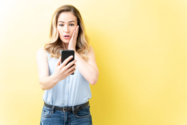Geschockte erwachsene Frau blickt auf Handy, während sie vor gelbem Hintergrund steht - Foto, Bild