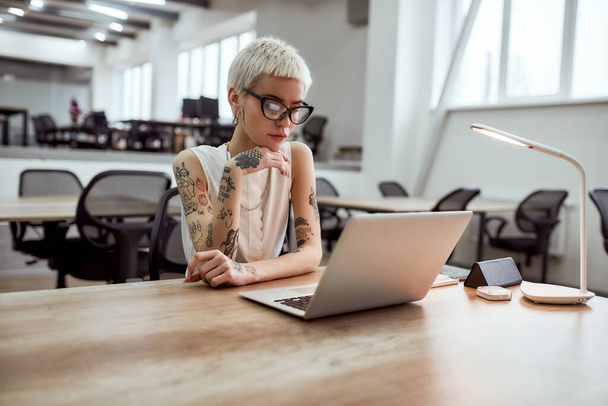 Busy day. Portrait of focused blonde tattooed businesswoman in eyeglasses looking at laptop screen while working in the modern working space - Foto, Imagem