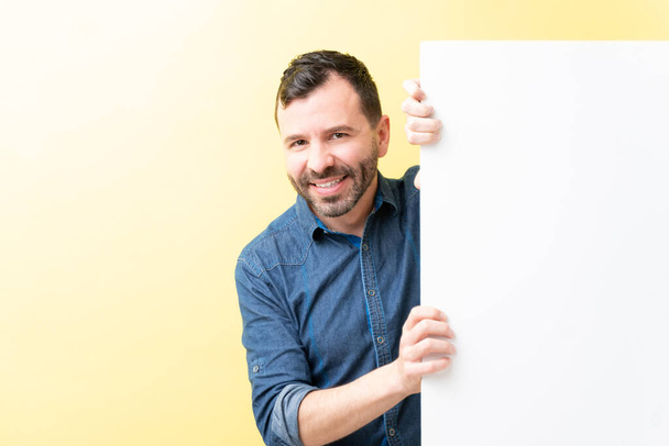 Bonito homem adulto escondido atrás de cartaz em branco contra fundo amarelo
 - Foto, Imagem