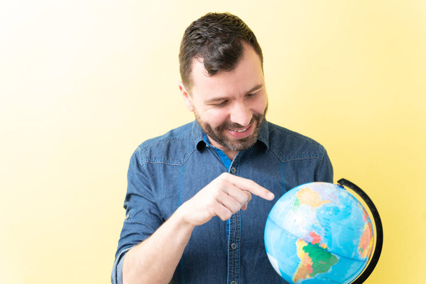 Smiling Latin man pointing at globe against yellow background - Foto, Bild