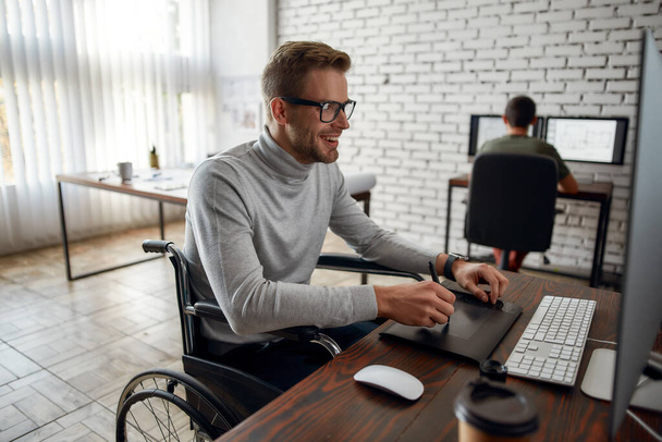 Enjoying his work. Side view of young and positive male office worker in a wheelchair using stylus pen and digital tablet while working in co-working space - Photo, Image