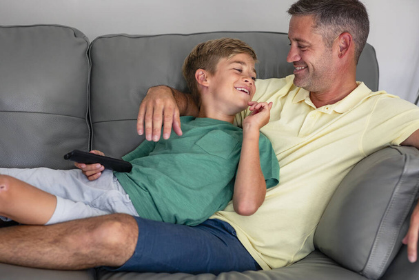 A shot of a young boy lying down on a couch watching tv with his dad, they are wearing casual clothing. - Фото, изображение