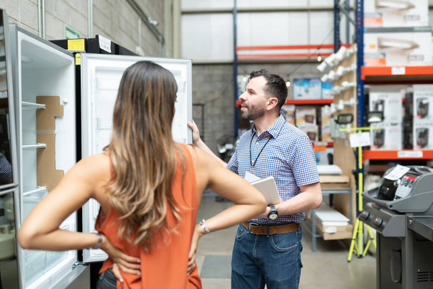 Male employee showing while explaining about refrigerator to female customer standing at warehouse store - Photo, Image