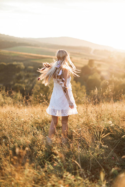 Young hipster girl in white dress and hair feathers standing outdoors, in summer field in mountains. Boho hippie style - Foto, immagini