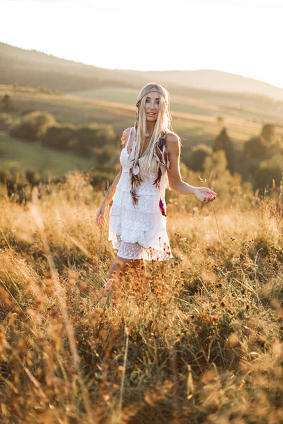Portrait of happy smiling woman with long blonde hair is wearing white boho hippie dress in field. Feathers in hair, boho style - Photo, Image