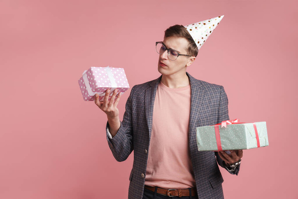 Image of a young thinking serious guy in glasses holding birthday present gift boxes isolated over pink wall background. - Фото, зображення