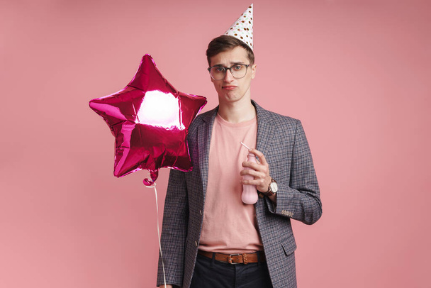 Image of a sad young boy in glasses in birthday cap isolated over pink wall background holding star balloon. - Фото, зображення