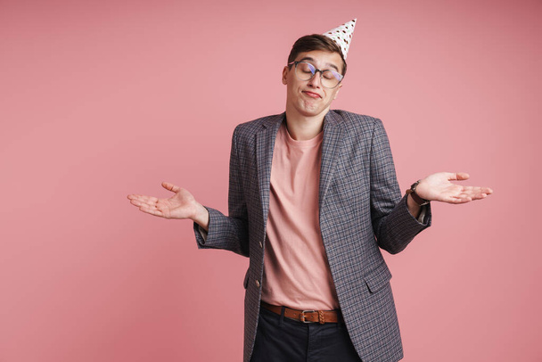 Portrait of an upset young man wearing birthday hat celebrating isolated over pink background, shrugging shoulders - Foto, Imagem