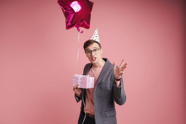 Portrait of a confused young man wearing birthday hat celebrating isolated over pink background, holding present box and balloon - Fotó, kép