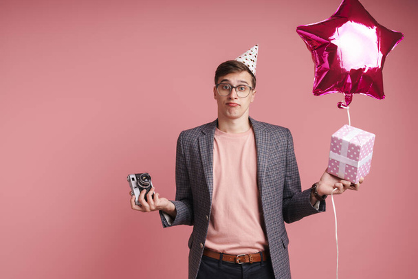 Porträt eines aufgebrachten jungen Mannes mit Geburtstagsmütze, der isoliert auf rosa Hintergrund feiert, Geschenkschachtel und Luftballon in der Hand, Fotokamera - Foto, Bild