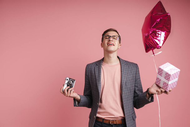 Portrait of an upset young man wearing birthday hat celebrating isolated over pink background, holding present box and balloon, holding photo camera - Foto, immagini