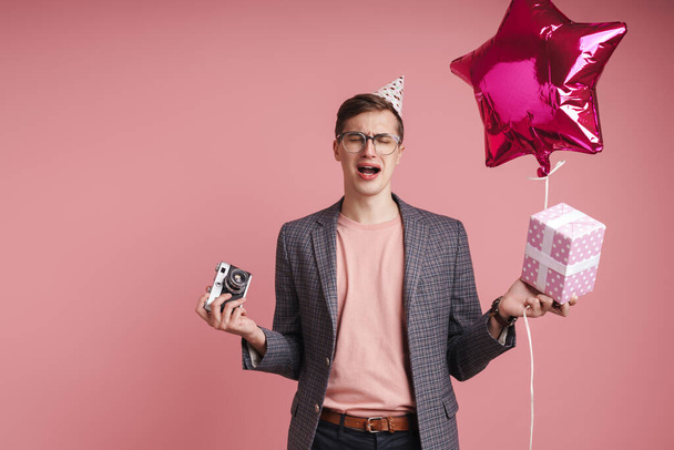 Portret van een overstuur jongeman met een verjaardagshoed die geisoleerd viert over een roze achtergrond, een doos en ballon vasthoudt, een fotocamera vasthoudt - Foto, afbeelding