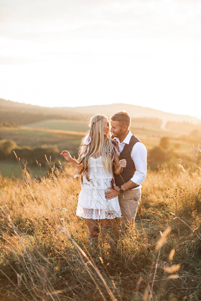 Beautiful couple in love embracing, at the sunset in summer field. Girl in white summer dress and feathers in hair, man in stylish casual suit. Summer vacation concept, boho couple - Photo, image