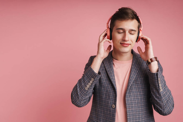 Portrait of a smiling young attractive man wearing jacket standing isolated over pink background, lstening to music with wireless headphones - Фото, зображення