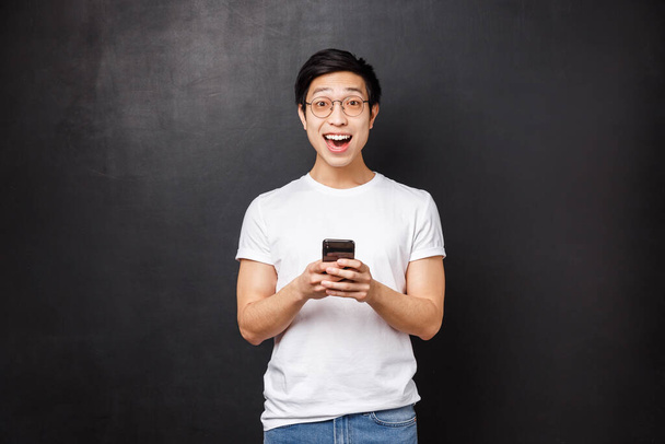 Tecnologia, gadgets e conceito de pessoas. Super feliz sorrindo positivo asiático cara em óculos e t-shirt, reagir divertido e maravilhado com notícias incríveis recebidas via e-mail, segure celular olhar câmera
 - Foto, Imagem