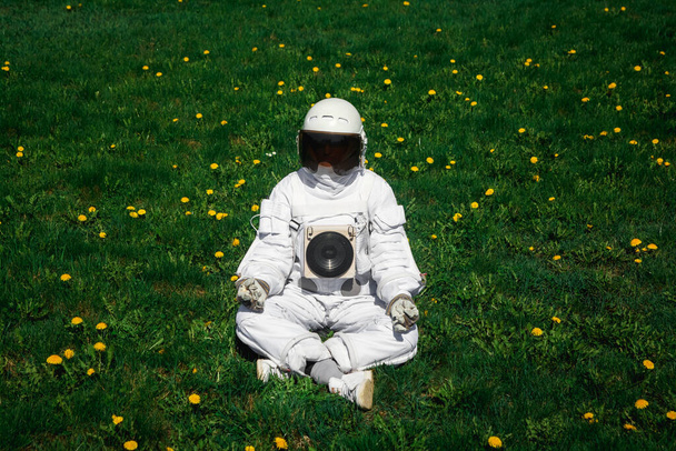 ヘルメットの未来型宇宙飛行士は花の間に緑の芝生の上に座っています。カメラへようこそ - 写真・画像