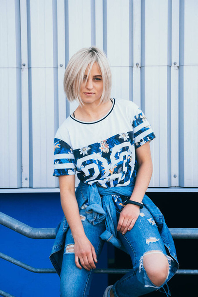 Beautiful girl with short white hair dressed in jeans in urban style stands on the background of a white wall. Her hair cover half face. She is looking forward. A look at the camera - Photo, Image