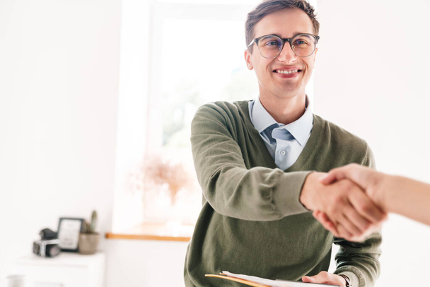 Image of a happy young guy indoor in office wearing eyeglasses shaking hand of his colleague. - Photo, image