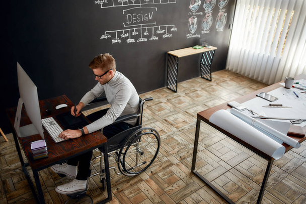 Professional at work. Young and creative male office worker in a wheelchair using stylus pen and digital tablet while sitting at his workplace in the modern office - Foto, Bild